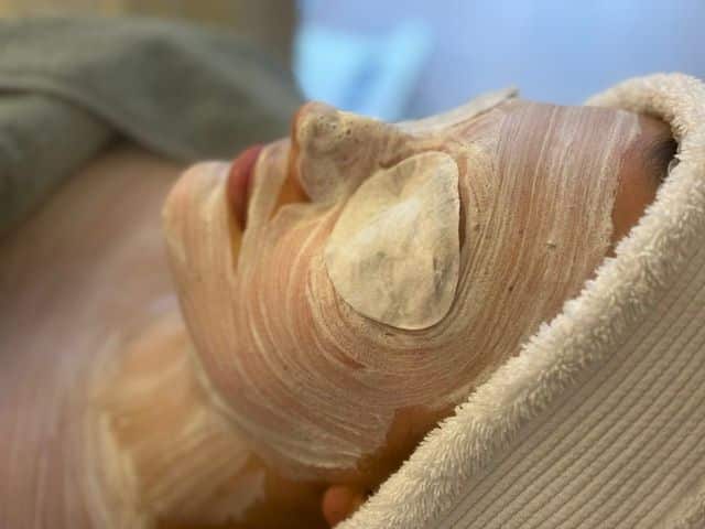 Klant met een verzorgend masker op het gezicht, ontspannen onder een handdoek in de behandelstoel.