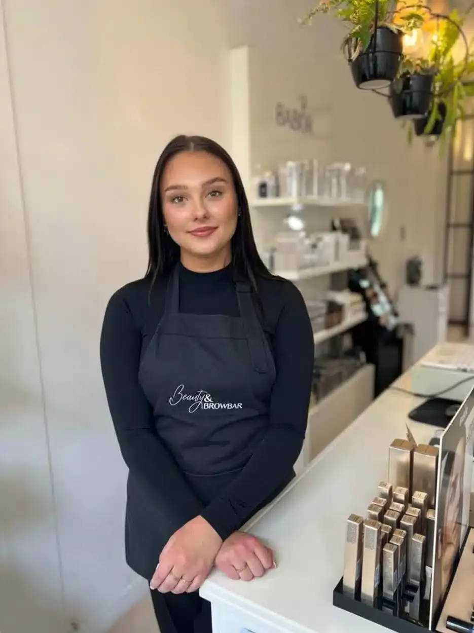 Senna staat aan de balie van de salon met het schort van Beauty & Browbar voor.